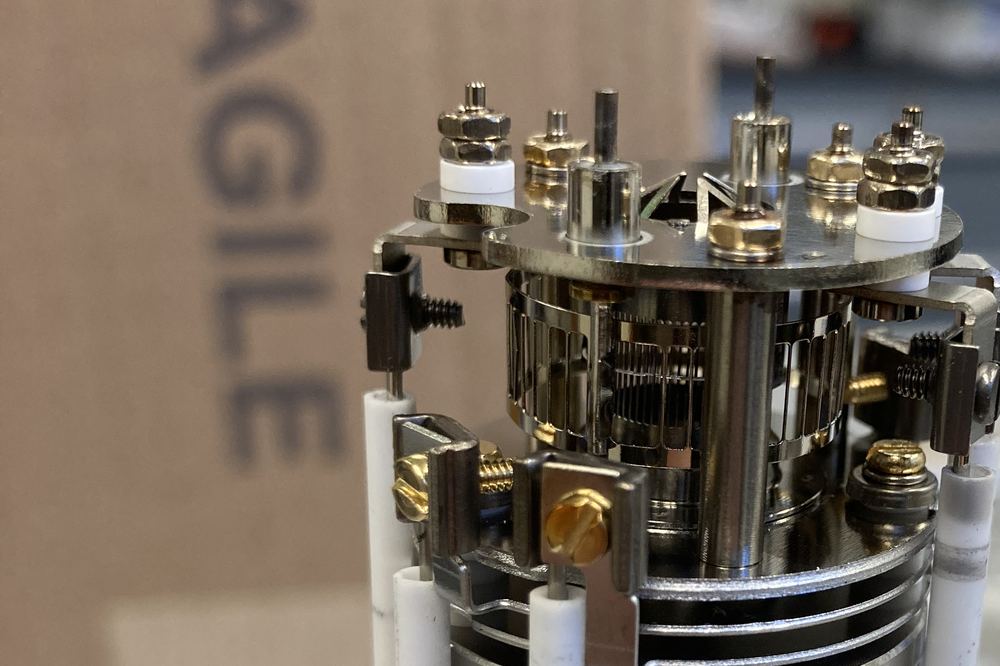 Close-up of a mass spectrometer ion source on a lab bench: a steel housing ringed by ceramic rods and gold-plated screws.